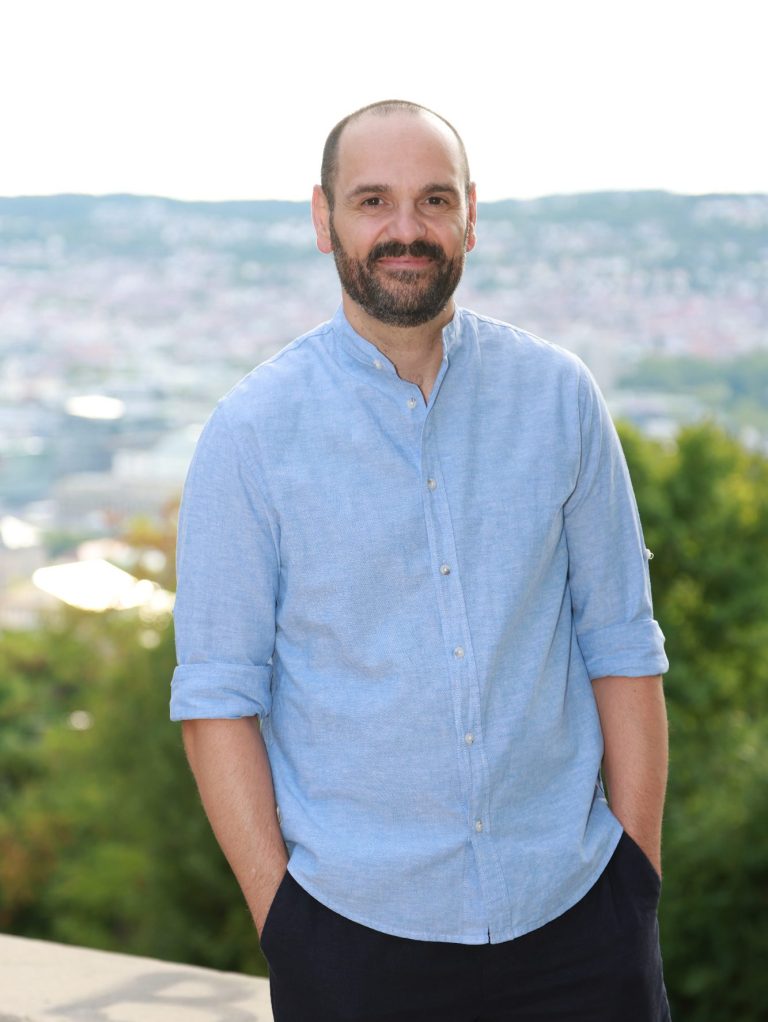 Claudio Capasso im hellblauen Hemd, lacht sympathisch und lehnt an einem Baumstamm mit locker verschränkten Armen an.