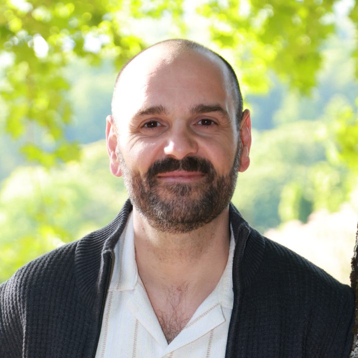 Claudio Capasso mit kurzem Bart, lächelnd vor einer grünen Landschaft.